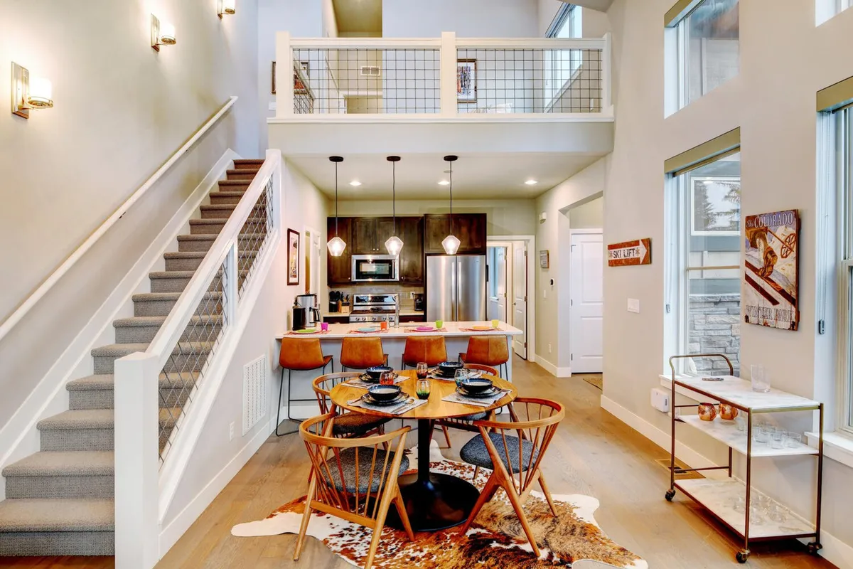 An open-plan interior scene where a spacious dining table, kitchen area, and large windows create a bright and welcoming atmosphere. The space is set for group meals and relaxation, featuring comfortable seating and modern design elements.