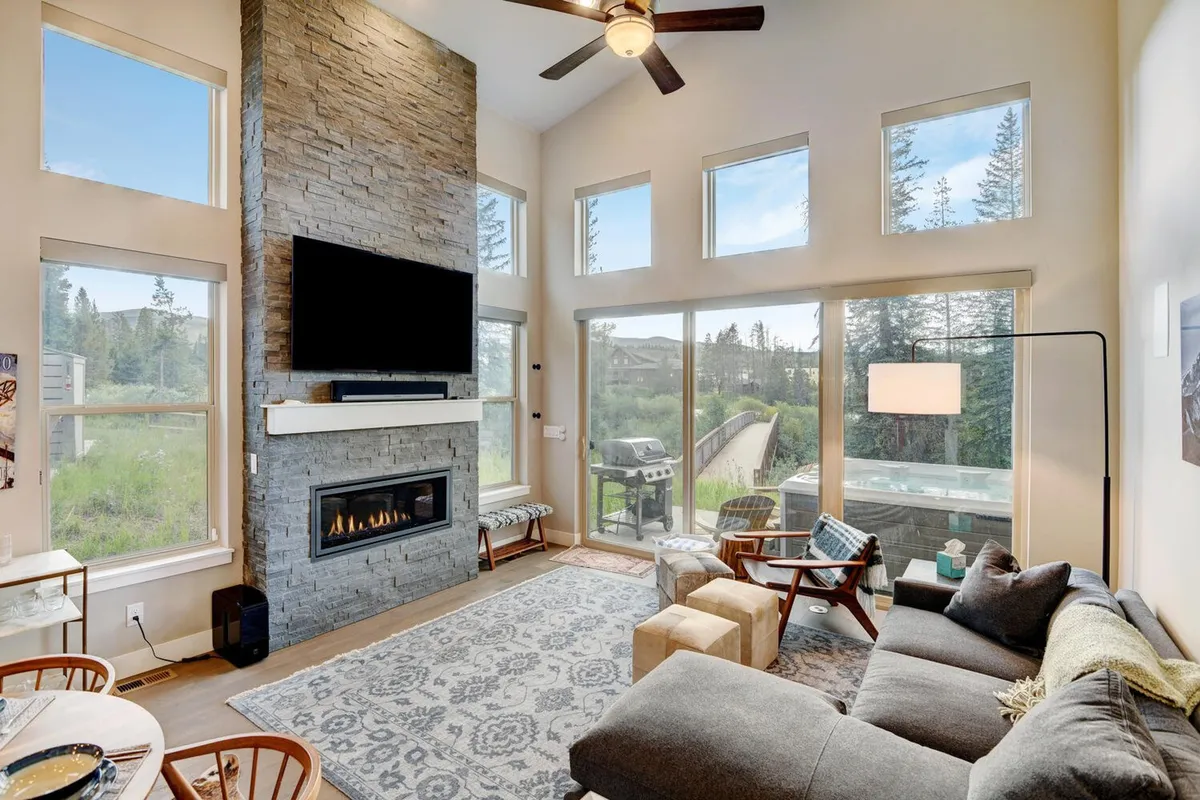 The image showcases a warm, inviting living room with a striking stone fireplace as the centerpiece. Large windows frame breathtaking sunrise mountain views, while plush seating invites relaxation. The open layout features a television and ample space for entertaining, enveloped in natural light and a cozy, mountain-cabin ambiance.
