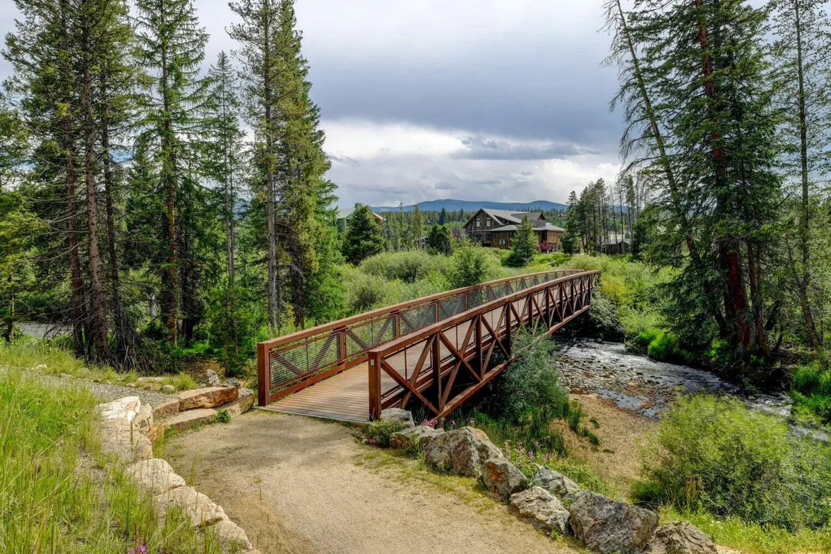 A tranquil scene featuring a wooden bridge crossing a stream, surrounded by lush trees and a dramatic mountain backdrop. The house is visible in the distance, suggesting proximity to nature trails and scenic walks. The peaceful setting invites both people and pets to explore and enjoy the outdoors.