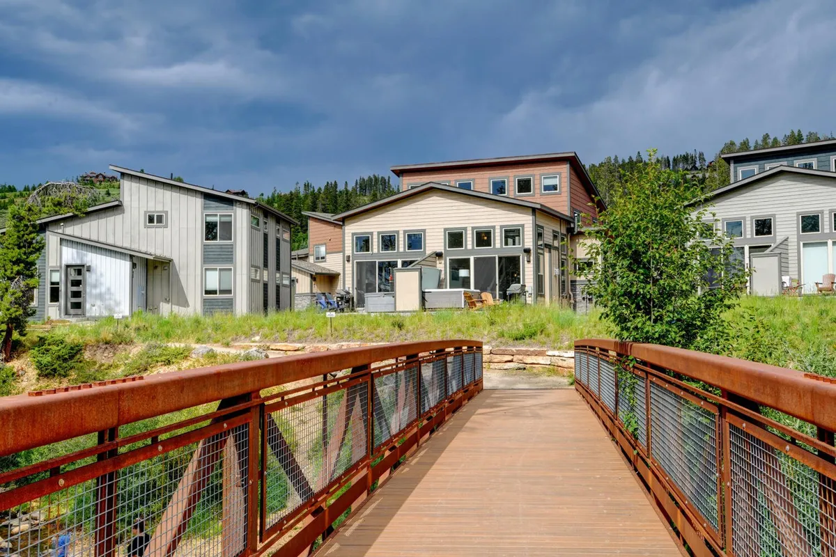 A tranquil scene featuring a charming wooden bridge, manicured green landscaping, and glimpses of modern houses in the background. The lush environment and welcoming outdoor spaces evoke a resort-like ambiance that connects the home to its scenic surroundings.