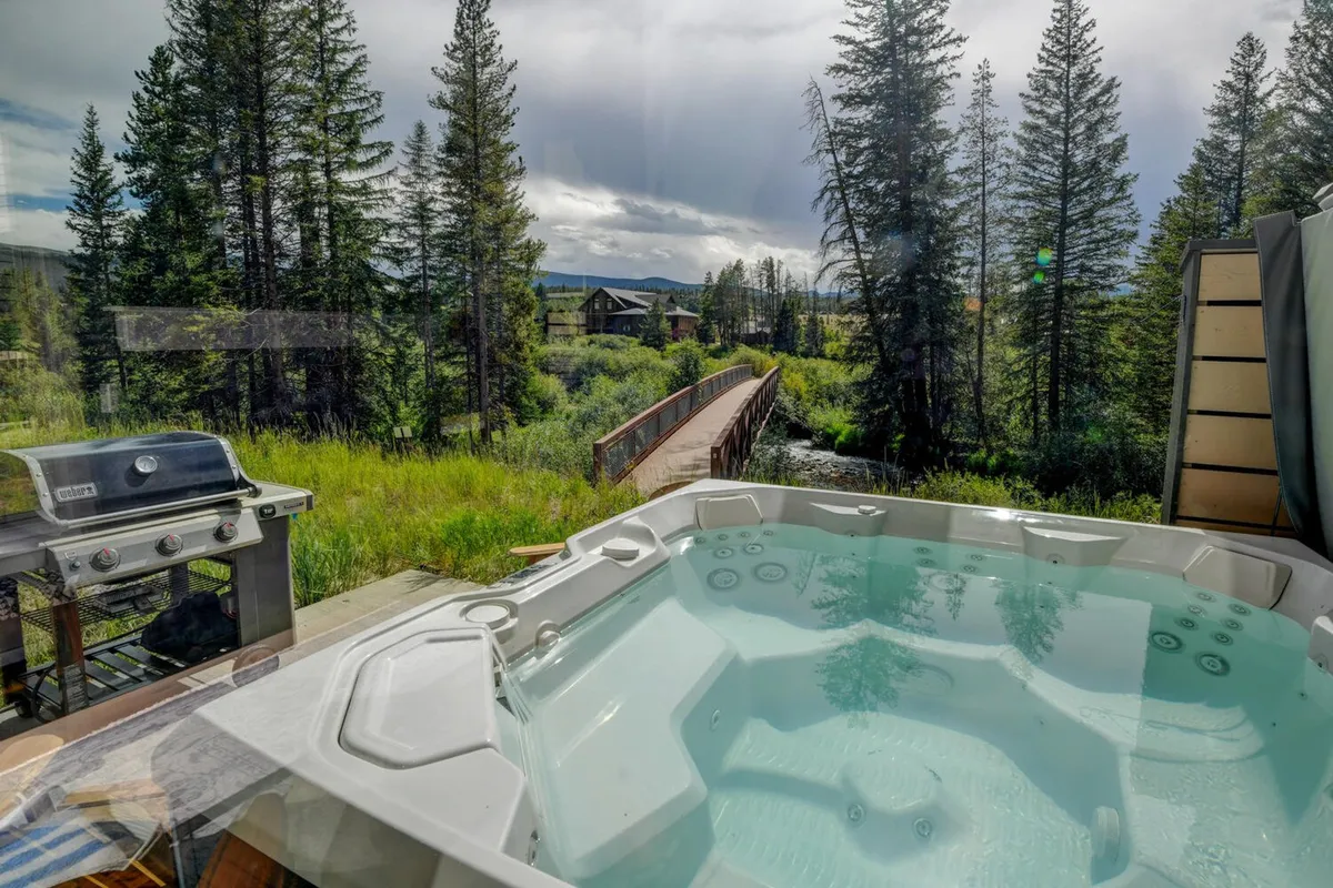 The photo highlights a hot tub on a wooden deck, adjacent to a grill and a wooden bridge, all set amidst lush trees. The peacefulness of the area is emphasized, perfect for both relaxation and outdoor adventures.