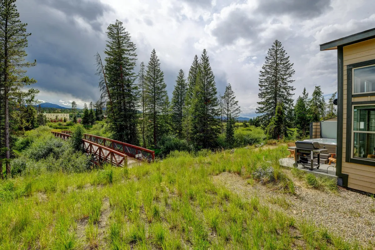 A serene outdoor space with a wooden bridge, dense trees, a mountain view, and a visible patio area. The scene evokes a sense of relaxation and adventure, inviting residents and their pets to enjoy both restful and active moments in nature.
