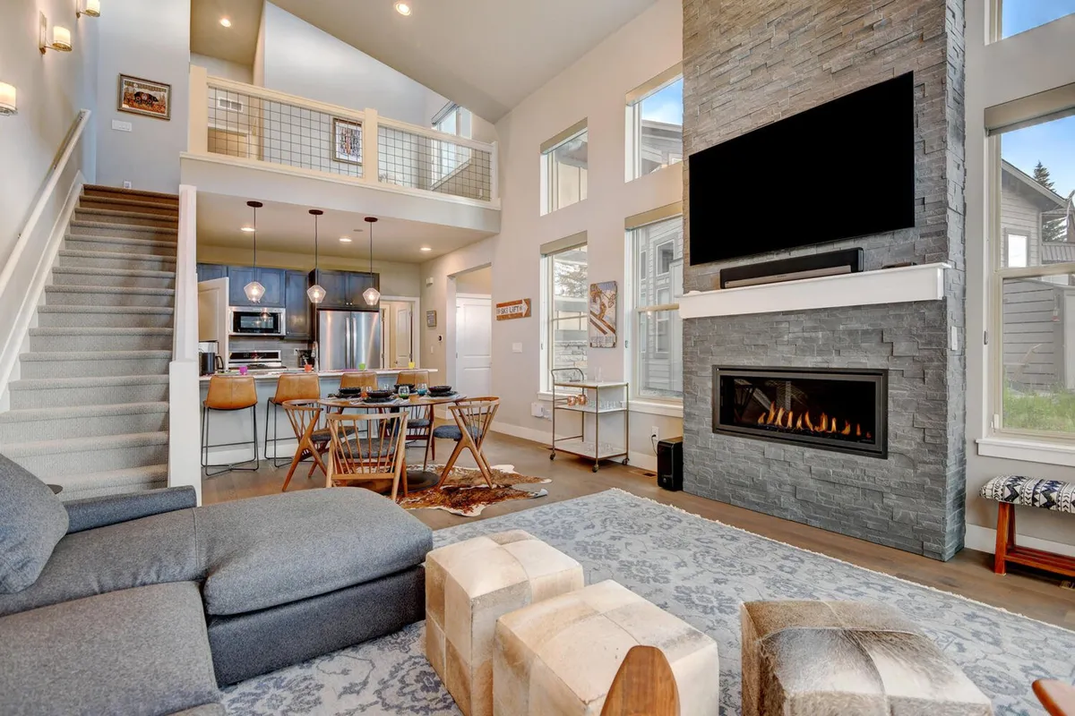 The photo features a plush sofa arranged around a striking stone gas fireplace, with a dining table and staircase in view. Large windows let in abundant natural light, enhancing the cozy, inviting atmosphere ideal for relaxing and connecting.