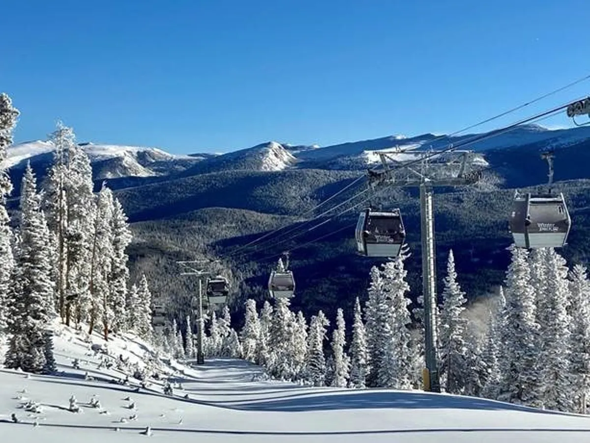 A panoramic exterior shot of a ski lift gliding above snow-covered trees, with majestic mountain peaks and a vibrant blue sky in the background. People can be seen enjoying skiing and snowboarding, adding to the scene's adventurous, active atmosphere.